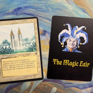 Peut inclure: Une carte à collectionner Magic: The Gathering intitulée The Tabernacle at Pendrell Vale avec une illustration de cathédrale et du texte, à côté d'une carte noire avec une illustration de bouffon et le texte jaune The Magic Lair.