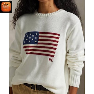 May include: A white long-sleeve crewneck sweater featuring a large knitted American flag graphic on the front. The flag design includes a blue field with white stars and red and white stripes. The letters RL are embroidered in red below the flag.