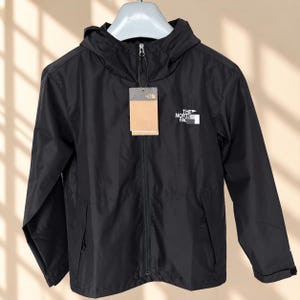 May include: A black hooded windbreaker jacket featuring a full-length front zipper, long sleeves with adjustable cuffs, and two side pockets. The North Face logo is printed in white on the chest, and a brown cardboard brand tag is attached.