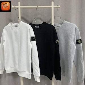 Op de afbeelding: Drie sweatshirts met lange mouwen en ronde hals in wit, zwart en gemêleerd grijs, elk voorzien van een cirkelvormige geborduurde patch op de linkermouw. De kledingstukken hangen aan individuele kledinghangers aan een horizontaal rek.