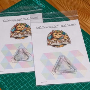 May include: Two clear acrylic quilting templates in triangular shapes, labelled 1 inch and 1.5 inch with 60 degree angles and 1/4 inch seam allowances. Each template is packaged in a plastic sleeve with a Ziggy Wiggles logo featuring a cat illustration.