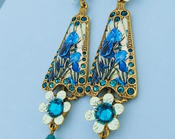 Puede incluir: Un par de pendientes colgantes dorados con un panel central triangular que presenta un estampado floral de iris azules. El diseño incluye detalles de cristales azules brillantes, elementos metálicos en forma de flor blanca y cuentas en forma de lágrima.