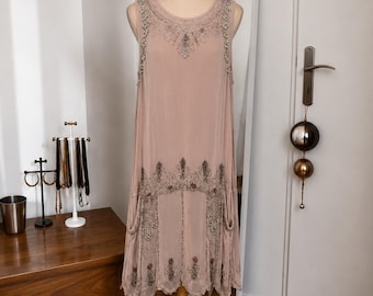 May include: A sleeveless, knee-length dress in a dusty rose hue, featuring intricate silver and dark grey beadwork along the neckline, shoulders, and lower hem. The garment has a relaxed, straight silhouette with a scalloped edge at the bottom.