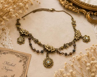 Può includere: Una collana in metallo color ottone con motivi floreali, ciondoli a forma di fiore pendenti e piccole perline scure sfaccettate. L'elemento centrale è un ciondolo a fiore circolare con una pietra iridescente al centro.