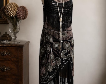 May include: A black sleeveless dress featuring thin straps, intricate silver and black sequin embroidery in a floral pattern, and a layered fringe hemline. A long, multi-strand white pearl necklace is draped over the bodice of the dress.