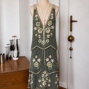 Puede incluir: Un vestido sin mangas hasta la rodilla en color verde oliva oscuro con falda de capas escalonadas y escote en V profundo. La prenda está ricamente adornada con abalorios en un patrón hexagonal y motivos florales en crema y verde.
