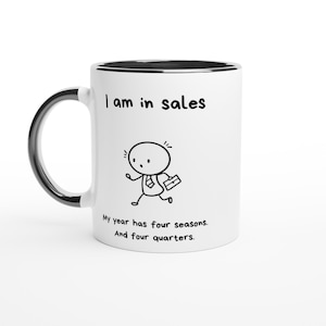 Peut inclure: Un mug en céramique blanche avec un intérieur et une anse noirs. L'avant présente un dessin au trait noir d'une personne avec une mallette et le texte : I am in sales. My year has four seasons. And four quarters. Le motif est centré.