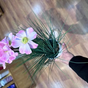 Könnte beinhalten: Eine künstliche Topfpflanze mit mehreren hellrosa und weißen Blüten mit gelben Mitten, begleitet von langen, dünnen, dunkelgrünen grasartigen Halmen. Die Pflanze befindet sich in einem einfachen weißen zylindrischen Topf.