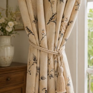 Puede incluir: Una cortina de color beige con un patrón estampado de pájaros, ramas y flores. La tela está fruncida y sujeta con un grueso alzapaños de cuerda trenzada que incluye una borla decorativa que cuelga del lateral del panel de la cortina.