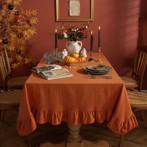 May include: A solid burnt orange rectangular tablecloth with a ruffled edge draped over a dining table. The table is decorated with a white vase, small pumpkins, two black spiral candles in gold holders, a book, and small ghost figurines.