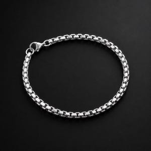 Op de afbeelding: Een zilverkleurige box chain armband bestaande uit een reeks onderling verbonden vierkante schakels die een doorlopende, flexibele band vormen. De armband is voorzien van een gepolijste karabijnsluiting met een uniforme metalen afwerking.