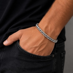 Op de afbeelding: Een zilverkleurige tarwekettingarmband met een gepolijste metalen afwerking en een geweven, in elkaar grijpend schakelontwerp. De armband is opgebouwd uit een reeks verbonden, afgeronde metalen schakels die een getextureerd uiterlijk creëren.