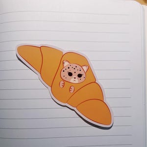 May include: A die-cut sticker featuring a golden-brown croissant with a cartoon leopard face peeking out from the center. The leopard has dark spots, large dark eyes, and a small heart-shaped nose, with two small paws visible on the pastry surface.