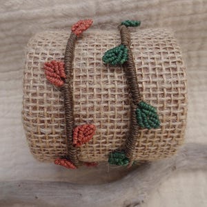 May include: Two macrame bracelets wrapped around a burlap roll. One bracelet features brown cord with small, knotted rust-colored leaf shapes. The second bracelet features brown cord with small, knotted dark green leaf shapes along the length.