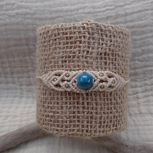 May include: A macrame bracelet featuring a central round blue stone bead surrounded by intricate beige knotted cord patterns and small metallic gold-toned beads. The bracelet is displayed wrapped around a textured tan burlap cylinder.