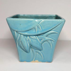 May include: A square-shaped ceramic planter with a glossy light blue glaze. The exterior features a raised, molded relief pattern of stylized leaves and stems that wrap around the sides of the vessel, creating a textured surface finish.