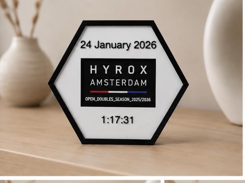 Op de afbeelding: Een zeshoekig wit bord met een zwarte rand met de tekst 24 January 2026, HYROX AMSTERDAM, een rode, witte en blauwe horizontale lijn, de tekst OPEN_DOUBLES_SEASON_2025/2026 en de numerieke tijd 1:17:31 in zwarte verhoogde letters.