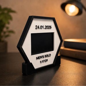 Op de afbeelding: Een zeshoekig zwart-wit frame met een centrale rechthoekige opening. Het witte oppervlak toont de tekst 24.01.2026, MEN'S SOLO en 1:17:31 in verhoogde zwarte letters. Het frame wordt ondersteund door een zwarte voet.