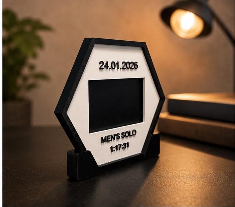 Op de afbeelding: Een zeshoekig zwart-wit frame met een centrale rechthoekige opening. Het witte oppervlak toont de tekst 24.01.2026, MEN'S SOLO en 1:17:31 in verhoogde zwarte letters. Het frame wordt ondersteund door een zwarte voet.