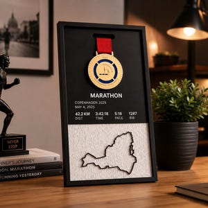 May include: A black rectangular display frame featuring a gold marathon medal with a red ribbon. The frame displays the text MARATHON, COPENHAGEN 2025, MAY 4, 2025, 42.2 KM, 3:42:18, 5:16, and 1287. Below the text is a white textured map outline.