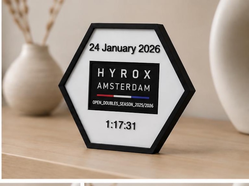 Op de afbeelding: Een zeshoekig zwart-wit displaybord met de tekst 24 January 2026, HYROX AMSTERDAM, een horizontale rood-wit-blauwe lijn, de tekst OPEN_DOUBLES_SEASON_2025/2026 en de numerieke tijd 1:17:31 in verhoogde zwarte letters.