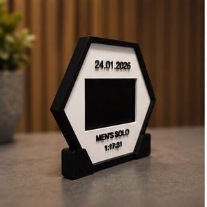 Op de afbeelding: Een zeshoekig zwart-wit frame met een centrale rechthoekige uitsparing. Het witte oppervlak toont de zwarte tekst 24.01.2026, MEN'S SOLO en 1:17:31. Het frame wordt ondersteund door een zwarte voet en heeft een strak geometrisch ontwerp.