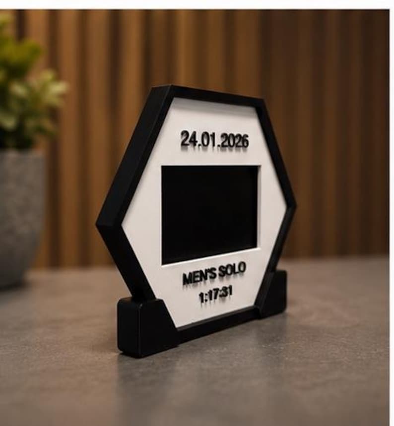 Op de afbeelding: Een zeshoekig zwart-wit frame met een centrale rechthoekige uitsparing. Het witte oppervlak toont de zwarte tekst 24.01.2026, MEN'S SOLO en 1:17:31. Het frame wordt ondersteund door een zwarte voet en heeft een strak geometrisch ontwerp.