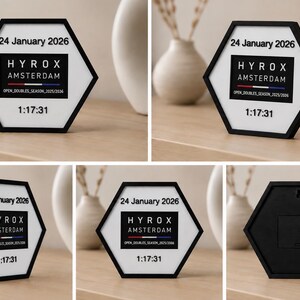 Op de afbeelding: Een zeshoekig displaybord met een zwarte rand en witte tekst met de tekst 24 January 2026, HYROX AMSTERDAM, OPEN_DOUBLES_SEASON_2025/2026 en 1:17:31. De achterkant van het frame is zwart met een ophanggat en een batterijcompartiment.