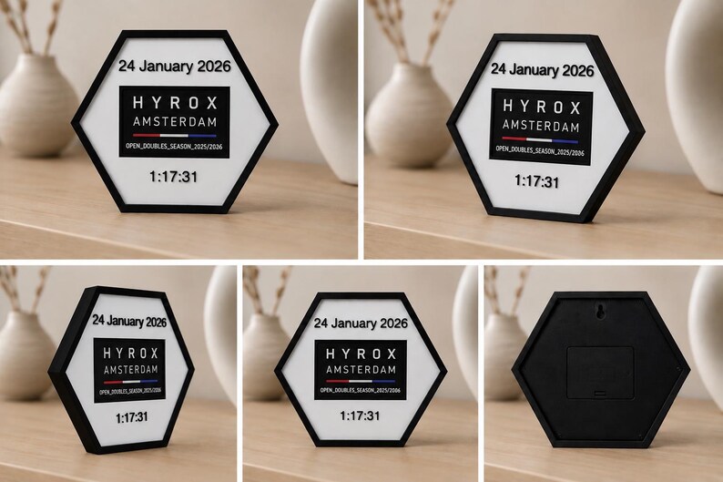 Op de afbeelding: Een zeshoekig displaybord met een zwarte rand en witte tekst met de tekst 24 January 2026, HYROX AMSTERDAM, OPEN_DOUBLES_SEASON_2025/2026 en 1:17:31. De achterkant van het frame is zwart met een ophanggat en een batterijcompartiment.
