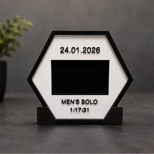 Op de afbeelding: Een zeshoekig zwart-wit displaybord met een centrale rechthoekige zwarte uitsparing. Het witte oppervlak toont bovenaan de datum 24.01.2026 en onderaan de tekst MEN'S SOLO gevolgd door 1:17:31 in verhoogde zwarte letters.