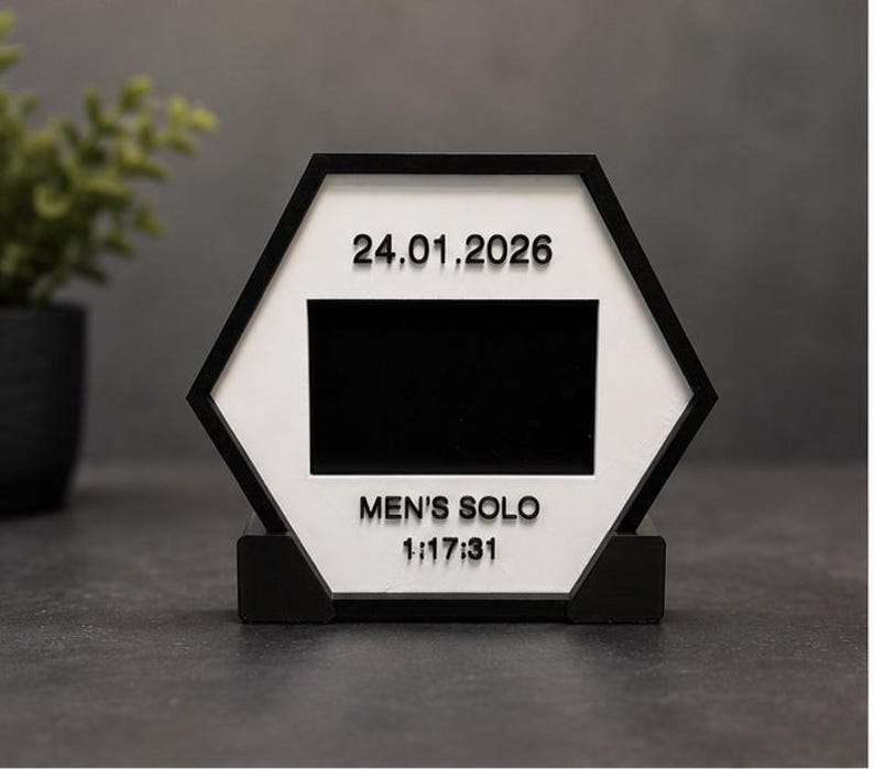 Op de afbeelding: Een zeshoekig zwart-wit displaybord met een centrale rechthoekige zwarte uitsparing. Het witte oppervlak toont bovenaan de datum 24.01.2026 en onderaan de tekst MEN'S SOLO gevolgd door 1:17:31 in verhoogde zwarte letters.