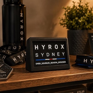 Può includere: Un espositore rettangolare nero con la scritta bianca HYROX SYDNEY, una linea orizzontale blu, bianca e rossa e il testo OPEN_DOUBLES_SEASON_2025/2026. L'espositore è posizionato vicino a una borraccia nera e a una medaglia metallica.