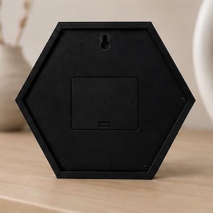 Op de afbeelding: Een zwarte zeshoekige klokachterkant met een centraal rechthoekig batterijcompartimentdeksel, een kleine sleutelgatvormige wandmontagesleuf in het midden bovenaan en zes kleine schroeven bij elke hoek van de rand van het frame.