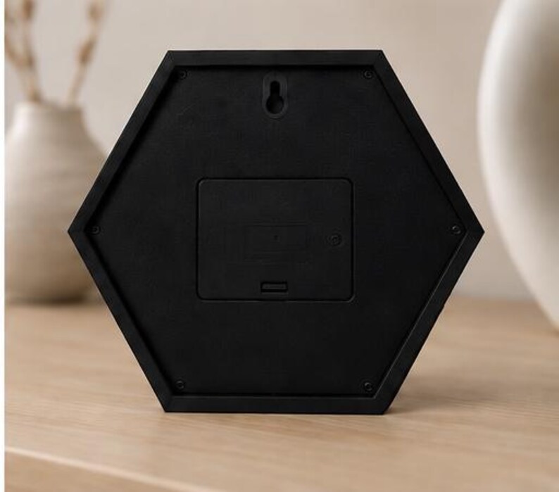 Op de afbeelding: Een zwarte zeshoekige klokachterkant met een centraal rechthoekig batterijcompartimentdeksel, een kleine sleutelgatvormige wandmontagesleuf in het midden bovenaan en zes kleine schroeven bij elke hoek van de rand van het frame.