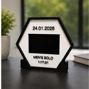 Op de afbeelding: Een zeshoekig zwart-wit displayframe met een centrale rechthoekige uitsparing. De witte voorzijde toont de tekst 24.01.2026 bovenaan en de tekst MEN'S SOLO 1:17:31 onderaan in verhoogde zwarte letters, rustend op een zwarte voet.