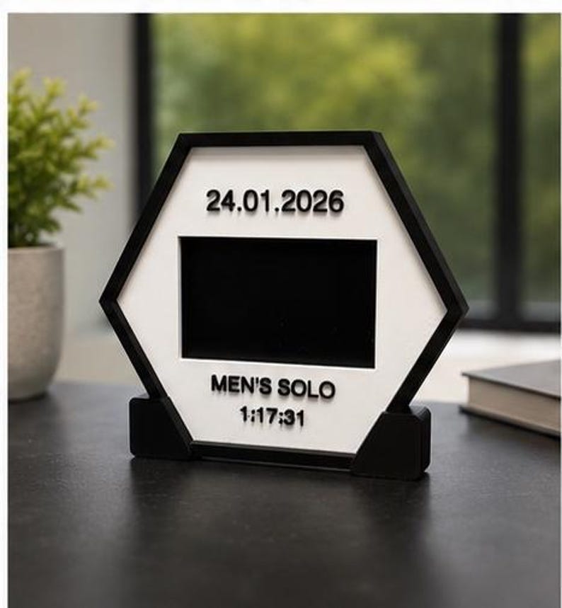 Op de afbeelding: Een zeshoekig zwart-wit displayframe met een centrale rechthoekige uitsparing. De witte voorzijde toont de tekst 24.01.2026 bovenaan en de tekst MEN'S SOLO 1:17:31 onderaan in verhoogde zwarte letters, rustend op een zwarte voet.