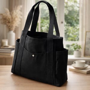 Op de afbeelding: Een effen zwarte tote bag met twee stevige verticale stoffen handvatten, een centraal buitenvak dat wordt gesloten met een zilverkleurige metalen drukknoop en twee open zijvakken, vervaardigd uit een duurzaam textielmateriaal.