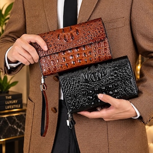 Può includere: Due pochette rettangolari con un motivo testurizzato in pelle di coccodrillo. Una pochette è marrone e l'altra è nera. Ogni pochette presenta una chiusura a patta e un cinturino da polso rimovibile fissato sul lato per un facile trasporto.