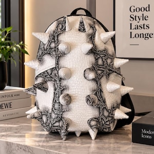 Puede incluir: Una mochila con estampado de piel de serpiente en blanco y negro, con grandes púas cónicas que sobresalen de la parte frontal. La bolsa tiene una parte superior redondeada, cierre de cremallera negro y un diseño estructurado con textura reptil.