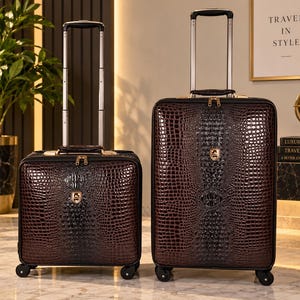 May include: Two dark brown suitcases with a textured crocodile skin pattern and a glossy finish. Each piece features a retractable silver handle, gold-tone zippers, and a small gold emblem on the front. Both suitcases stand on four black wheels.