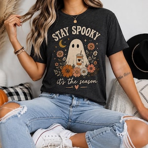 Pode incluir: Uma t-shirt de manga curta cinzento-carvão escuro com um gráfico de um fantasma a segurar um copo de café rodeado por flores, uma lua crescente e o texto "STAY SPOOKY" no topo e "it's the season" na parte inferior com um pequeno coração.
