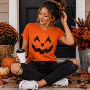 Pode incluir: Uma t-shirt laranja de manga curta com um gráfico de rosto de abóbora de Halloween em preto com um nariz em forma de coração, combinada com calças de ganga pretas rasgadas e ténis brancos. A t-shirt tem gola redonda e mangas dobradas.