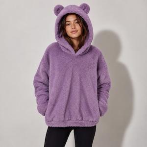 Könnte beinhalten: Ein lavendelfarbener Langarm-Kapuzenpullover aus weichem, flauschigem Kunstfellmaterial. Der Kapuzenpullover verfügt über eine Kapuze mit zwei kleinen, abgerundeten, bärenartigen Ohren an der Oberseite sowie eine große Kängurutasche auf der Vorderseite.