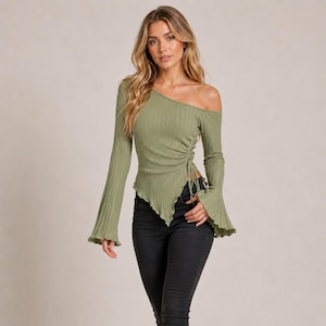 Può includere: Un top a maniche lunghe color verde oliva con texture a coste, caratterizzato da uno scollo asimmetrico a spalle scoperte, maniche a campana e un dettaglio con coulisse laterale che crea un effetto arricciato e un orlo a punta, abbinato a jeans neri.