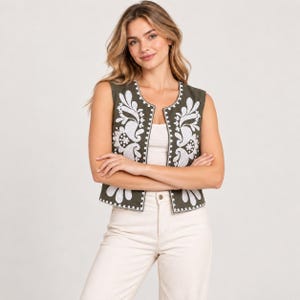 Puede incluir: Un chaleco sin mangas de color verde oliva con intrincados bordados florales blancos a lo largo de los paneles frontales y el dobladillo. El chaleco tiene un cuello redondo y se lleva sobre una camiseta blanca lisa con pantalones claros.