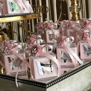 May include: A collection of square glass perfume bottles with ribbed pink labels, each featuring a silver butterfly cutout with the text Elisa Sengul. The bottles are adorned with a pink ribbon and a small fabric rose on the bottle neck.