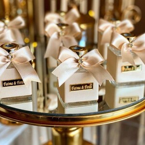 May include: A square glass bottle with a ribbed cream-colored texture, topped with a champagne-colored satin bow featuring a central rhinestone. A gold rectangular plaque on the front displays the black text Fatma & Ferdi in a serif font.