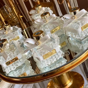 May include: A collection of square glass perfume bottles with gold-tone caps, each adorned with a white satin ribbon bow, a small rhinestone, and a white lace trim. A rectangular gold-tone plaque with the name Alya is attached to each bottle.