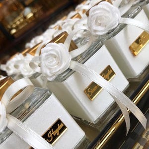 May include: A collection of square glass bottles featuring a white satin ribbon wrap, a white fabric rose accent, and a rectangular gold-toned metal plaque with the engraved text "Simon & Hayden" centered on the front of each bottle.