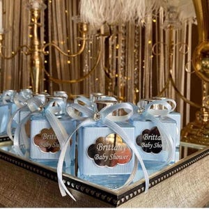 May include: A collection of square glass bottles filled with a light blue liquid, each adorned with a light blue ribbon bow and a cloud-shaped metallic label featuring the text Brittany Baby Shower in a decorative script font.